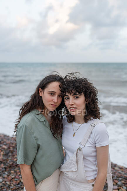 Jeunes copines lesbiennes en tenue décontractée embrassant tout en regardant la caméra sur la côte de l'océan sous un ciel nuageux — Photo de stock