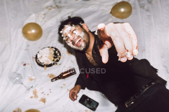 Drunk laughing male in smashed birthday cake lying near empty bottles from beer and balloons and pointing at camera — Stock Photo