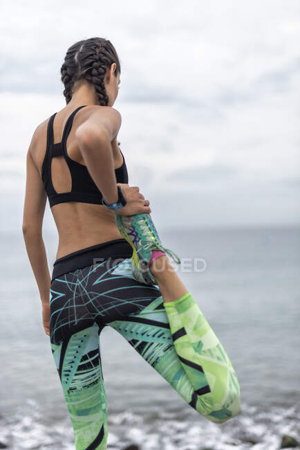 Back view of slim sportswoman stretching legs and waring up during workout on embankment in summer — Stock Photo