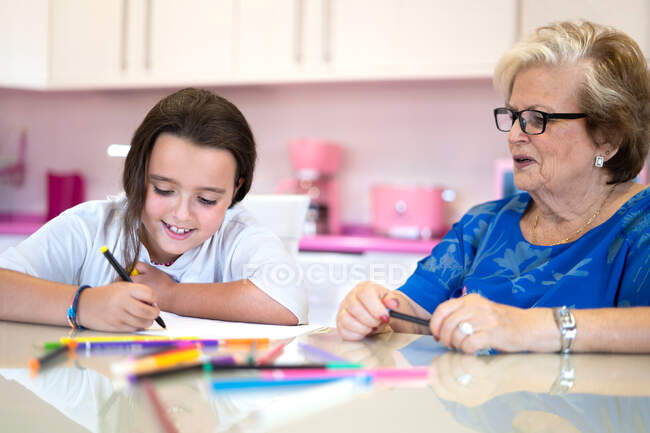 Улыбающаяся бабушка помогает веселой внучке рисовать на бумаге, проводя время вместе на кухне дома — стоковое фото