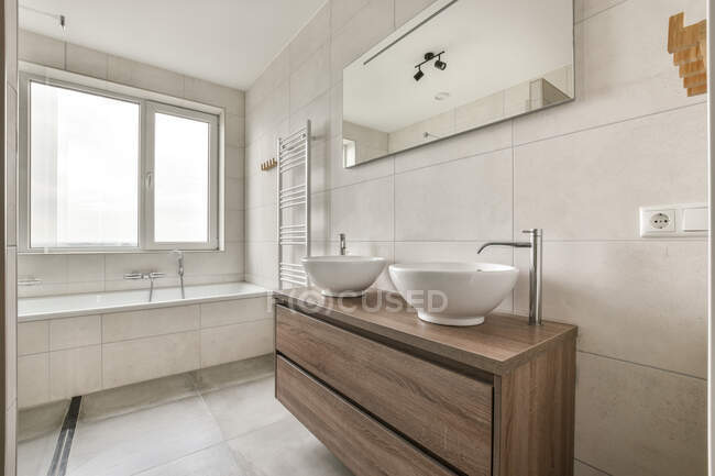 Двойная раковина на деревянном шкафу под зеркалом в светлой ванной комнате с ванной в современной квартире — стоковое фото