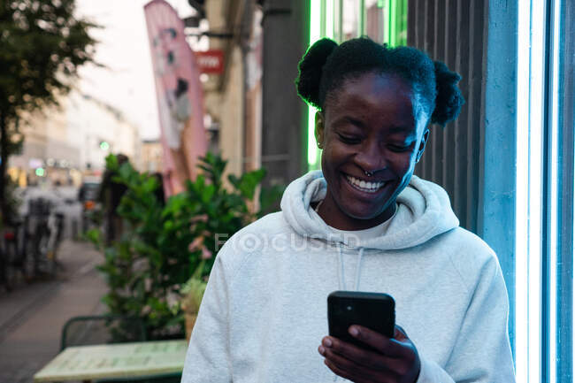 Positive Afroamerikanerin in lässigem grauen Kapuzenpulli am Handy, die auf den Bildschirm schaut, der auf der Straße in der Stadt läuft — Stockfoto