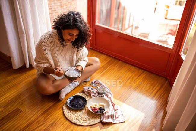 Alto ángulo de hembra sentada con las piernas cruzadas y comiendo plátanos en rodajas dulces arándanos y frambuesas - foto de stock