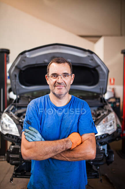 Позитивный зрелый мужчина в грязной рабочей одежде стоит со скрещенными руками на фоне автомобиля в ремонтной службе и смотрит в камеру — стоковое фото