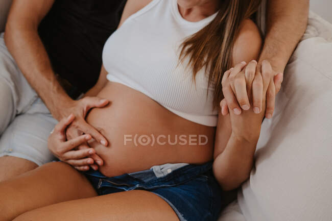 Cortado homem irreconhecível abraçando barriga de expectante fêmea amada enquanto descansa no sofá na sala de estar — Fotografia de Stock