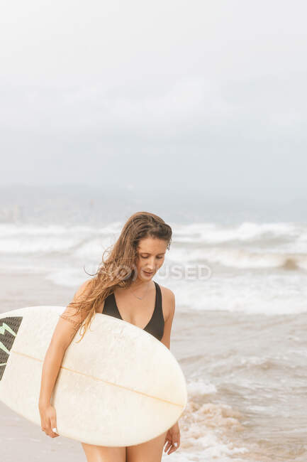 Задумчивая молодая спортсменка в купальниках с летящими волосами и доской для серфинга, смотрящая вниз на побережье океана — стоковое фото