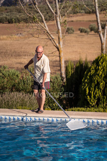 Cuerpo completo de hombre anciano en anteojos limpiando agua en piscina con fregona en patio trasero - foto de stock
