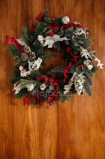 Elegante corona de Navidad con ramitas de coníferas y elementos decorativos colgando de la pared de madera a la luz del día - foto de stock