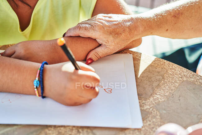 Зверху невідома бабуся торкається руки анонімної онуки, сидячи за столом і малюючи на папері в сонячний день — стокове фото