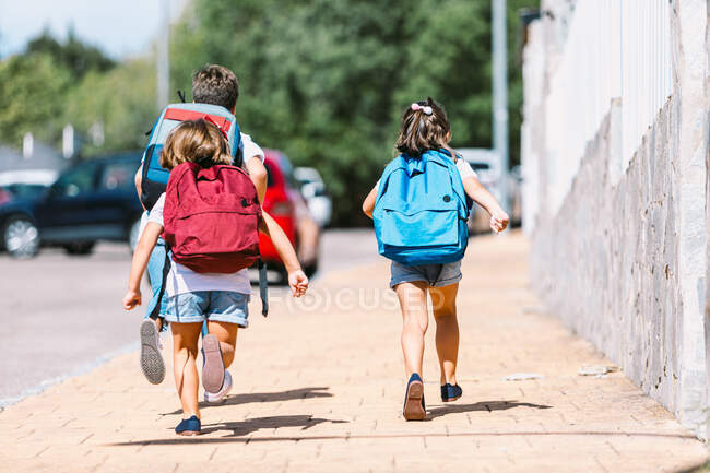 Vue arrière d'écoliers anonymes avec des sacs à dos circulant sur une passerelle carrelée dans une ville ensoleillée sur fond flou — Photo de stock