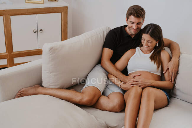 Hombre abrazando vientre de la mujer embarazada amada mientras descansa en el sofá en la sala de estar - foto de stock