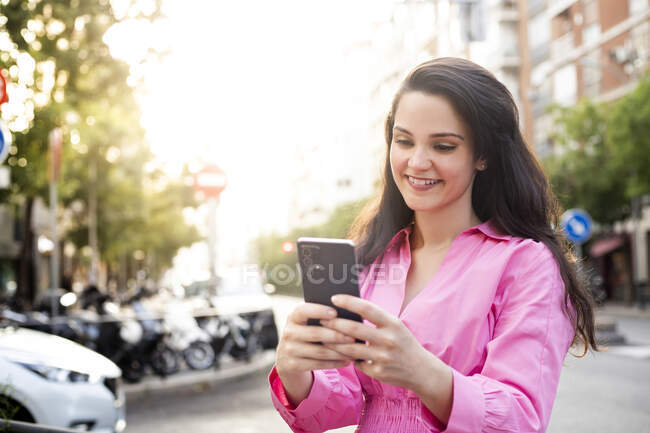 Donna sorridente in abito in piedi sul marciapiede e sms sul cellulare — Foto stock