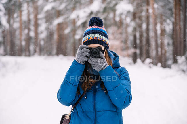 Молода жінка в теплому верхньому одязі стоїть серед сніжних дерев і фотографується на камеру в зимовому лісі — стокове фото