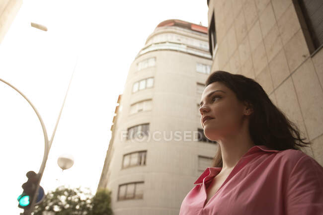 Низький кут серйозної жінки з довгим волоссям, що стоїть на тротуарі біля високої будівлі і світлофора — стокове фото