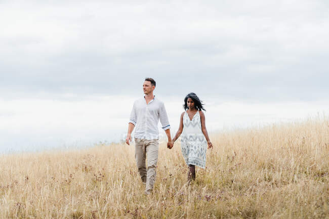 Мужчина с этнической девушкой, держащийся за руки во время прогулки по осеннему лугу под облачным небом — стоковое фото