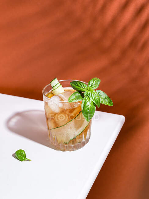 High angle of glass of cold cocktail with cucumber slice and basil leaves served on white table in red background — Stock Photo
