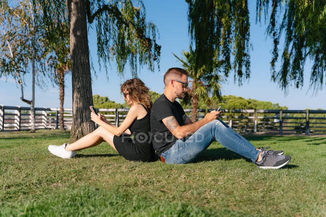 Side view of tattooed man with girlfriend in sunglasses browsing on cellphones while sitting back to back on meadow in summer park — Stock Photo