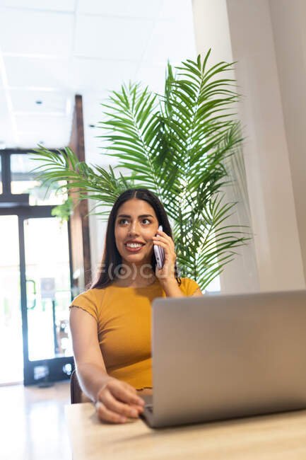 Щаслива фрілансерка сидить за дерев'яним столом у кафе і телефонує під час введення в мережу — стокове фото