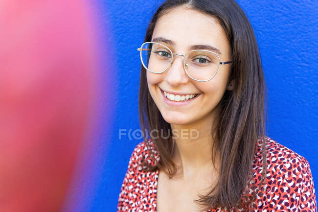 Femme heureuse en robe rouge élégante debout contre le mur bleu dans la journée en regardant la caméra — Photo de stock