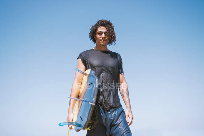 Young male athlete with curly hair and tattoos in wet sportswear holding surfboard while looking at camera under light sky — Stock Photo