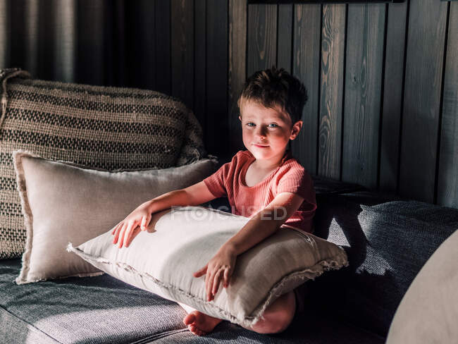 Contenuto adorabile bambino seduto su un morbido divano con cuscino in soggiorno e guardando la fotocamera — Foto stock