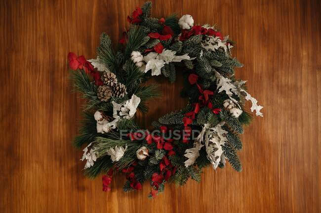 Elegante corona de Navidad con ramitas de coníferas y elementos decorativos colgando de la pared de madera a la luz del día - foto de stock