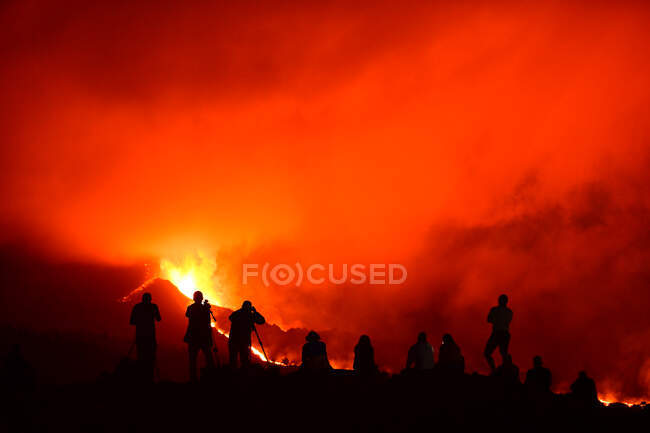 Силуэты человека, стоящие, записывающие и фотографирующие на штативах взрыв лавы на островах Пальма-Канарских островов 2021 года и несколько силуэтов, сидящих, наблюдая за природным явлением. — стоковое фото