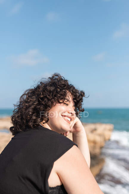 Весела молода товста самиця з закритими очима сидячи на грубій кам'янистій формації проти океану під хмарним небом на сонці. — стокове фото
