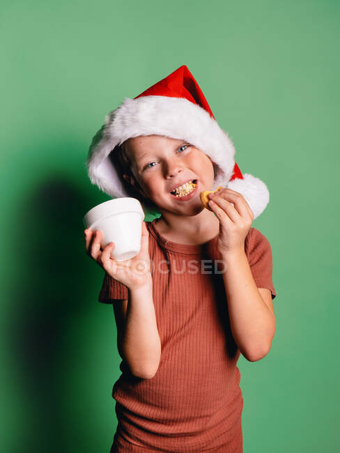 Чарівний усміхнений маленький хлопчик з різдвяним капелюхом Санта, беручи печиво з чашки на зеленому тлі, дивлячись на камеру — стокове фото