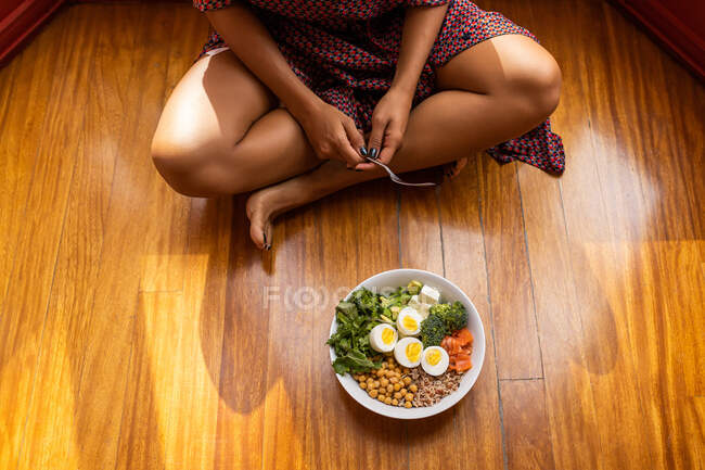 Angle élevé de la femelle récoltée méconnaissable assis avec les jambes croisées et manger super bol de nourriture avec pois chiche oeufs hachés brocoli — Photo de stock