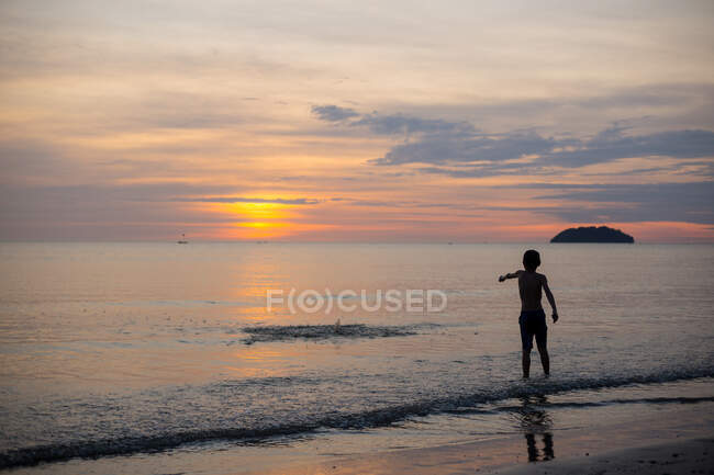 Back view full length of unrecognizable boy standing on wet sandy beach washed by sea at sundown in Malaysia — Stock Photo