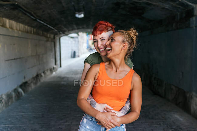Alegre joven mujer tatuada con el pelo rojo abrazando homosexual amado mientras se ríe en el túnel en la ciudad - foto de stock