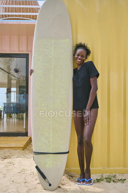 Cheerful young African American sportswoman in bikini and t shirt looking at camera with surfboard in a beach bar on the coast — Stock Photo