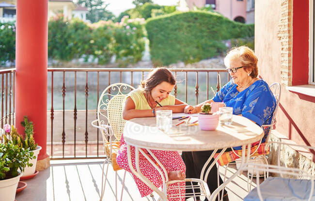 Улыбающаяся бабушка помогает веселой внучке рисовать на бумаге, проводя время вместе на террасе в солнечный день — стоковое фото