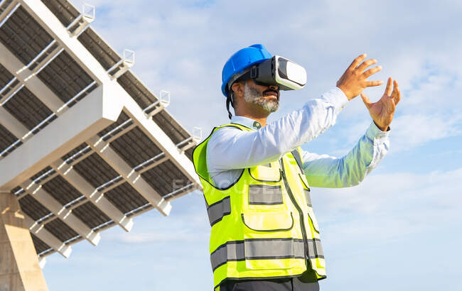 Low angle of bearded male inspector in uniform exploring virtual reality in headset against modern construction under cloudy sky — Stock Photo