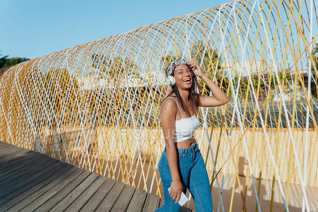 Mujer étnica joven contemplativa en bandana y auriculares inalámbricos escuchando canciones mientras se cierran los ojos en el puente urbano - foto de stock