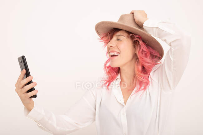 Slide view mujer alegre con pelo rosa en ropa elegante tomando selfie en el teléfono inteligente contra fondo claro - foto de stock