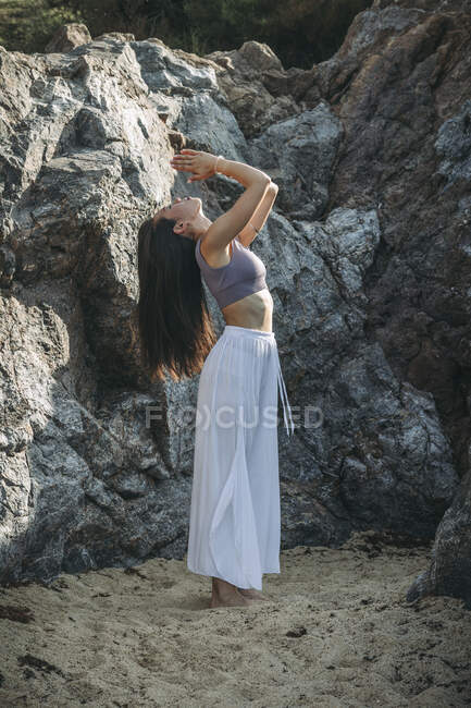 Side view of ethnic female with praying hands practicing yoga on sandy land against rock in sunlight — Stock Photo
