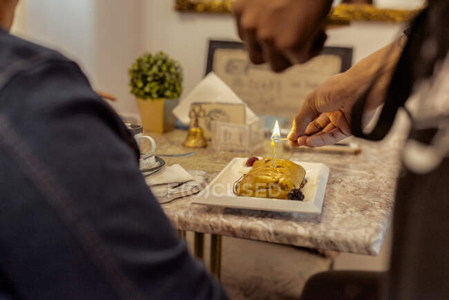 Crop unrecognizable cafe employee lighting birthday candle on tasty sweet pastry against client at table — Stock Photo