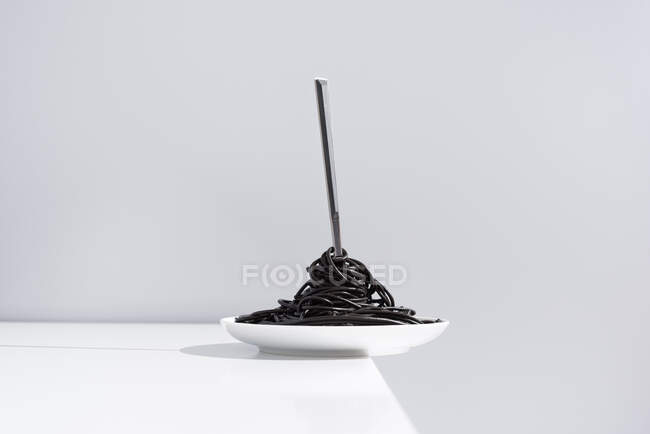Fourchette en acier inoxydable dans un bol plein de spaghettis noirs à l'encre de seiche sur table blanche en studio sur fond gris — Photo de stock