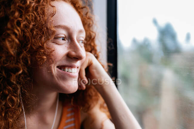 Cheerful young woman with curly red hair listening to music from earphones while looking away during trip on train — Stock Photo