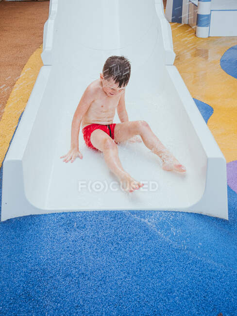 Full body of barefoot boy moving on wet slide under water drops in aqua park — Stock Photo