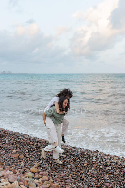 Cheerful young female giving lesbian girlfriend piggyback ride while having fun on pebble shore against wavy sea in twilight — Stock Photo