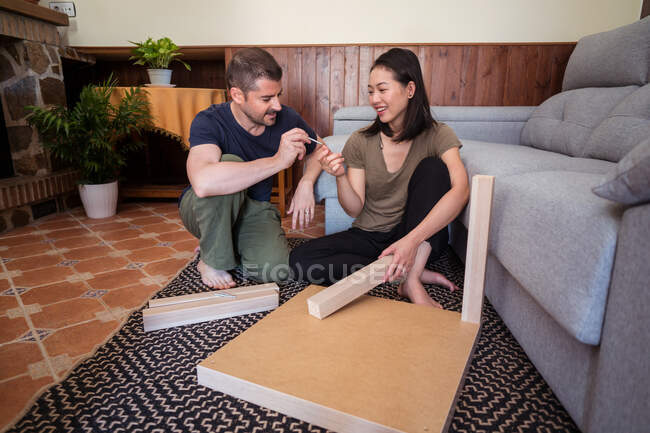Man passing screw to cheerful Asian wife while mounting table on ornamental carpet in living room — Stock Photo
