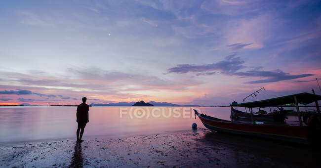 Полное тело силуэта неузнаваемого туриста, стоящего на мокром песчаном берегу возле лодки и любующегося морем под закатом неба в Малайзии — стоковое фото