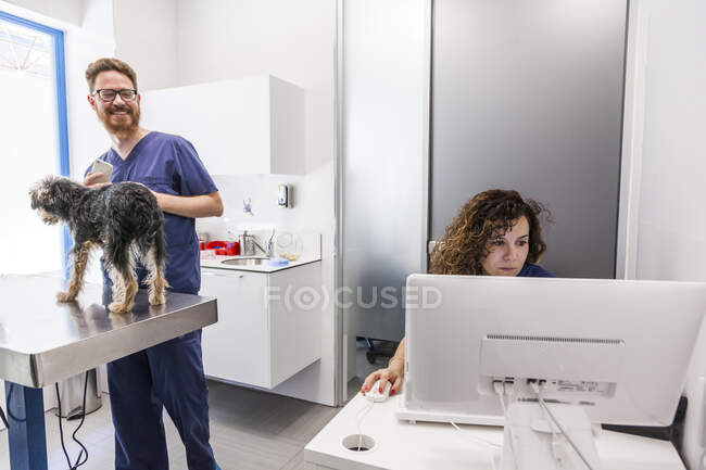 Sorrindo homem com smartphone acariciando fofo Yorkshire Terrier e falando com assistente do sexo feminino trabalhando no computador na clínica veterinária — Fotografia de Stock