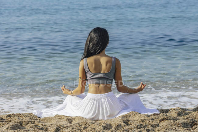 Rückansicht einer anonymen Frau, die in Padmasana-Pose sitzt, während sie Yoga an der Sandküste gegen das kräuselnde Meer praktiziert — Stockfoto
