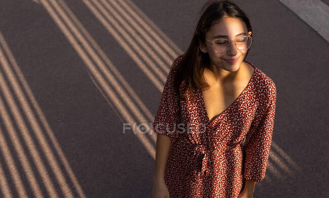 From above of cheerful female in dress and eyeglasses standing with eyes closed in sunlight — Stock Photo