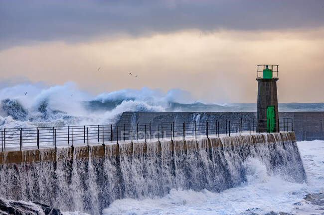 Огромные пенные морские волны врезаются в каменистый волнолом со старой башней маяка против голубого облачного неба в порту Виавлес в Астурии, Испания — стоковое фото