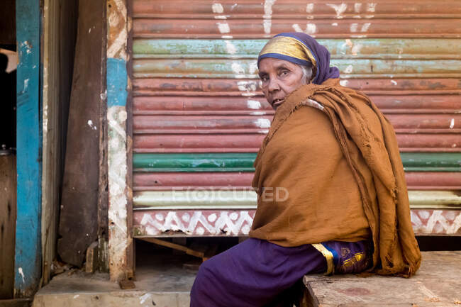 INDIA, BANGLADESH - 7 DE DICIEMBRE DE 2015: Mujer étnica mayor con ropa tradicional mirando a la cámara mientras está sentada cerca de un edificio en mal estado - foto de stock